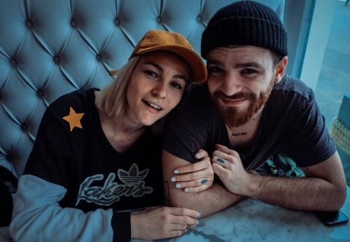 A photo of a young couple in a diner booth. They're tattooed, she's wearing a gold corduroy baseball hat and he's wearing a black beanie.