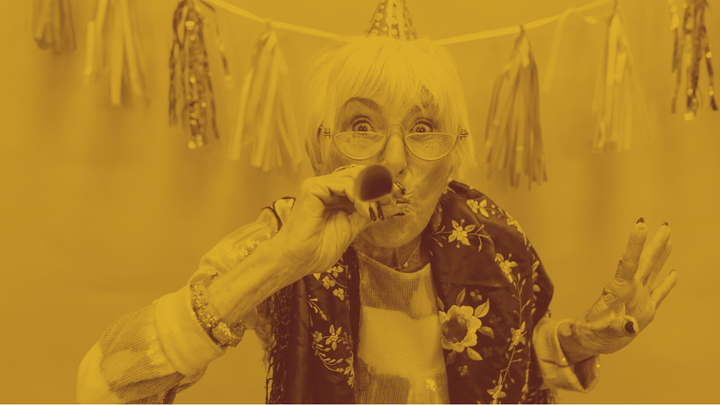 Photo of an old white woman with a party hat on blowing into a party horn.