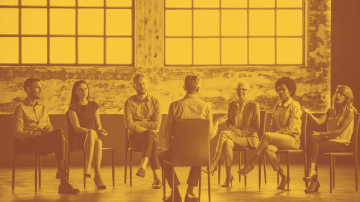 A photo of a group of people in chairs, 6 of them in a semi-circle facing one man