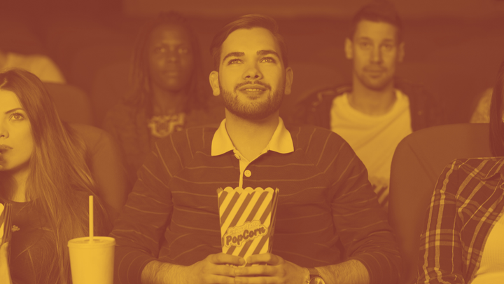 A photo of a young man grinning goofily, holding a box of popcorn while he stares at the movie screen