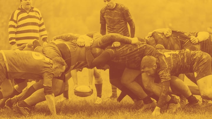 A photo of a bunch of male rugby players in a scrum, with a referee looking on.